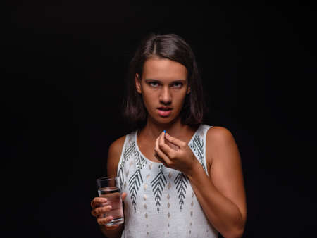 Young woman holding a glass of water and taking pills on a black background. Medical help, medicine concept. Copy space.の写真素材