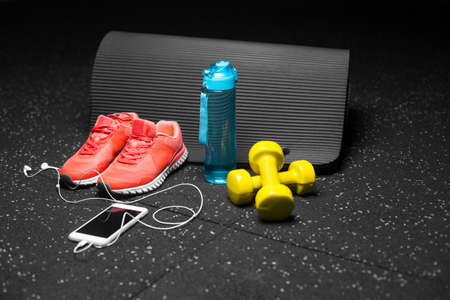 Small yellow dumbbells, pilates mat, blue bottle full of water, bright pink trainers and modern white smartphone with handphones on a black background. Sports and training concept. Copy space.の写真素材