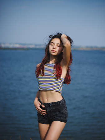 Sexy, cute young girl touching her hair. A teenager on a blurred river background. Beauty and style concept. Copy space.の写真素材
