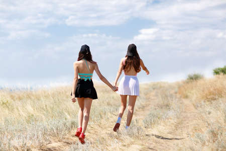 A picture of two beautiful, stunning young ladies walking hand in hand along the yellow field. Couple girls wearing colorful skirts and black baseball caps on a natural blue sky background. Copy pace.の写真素材