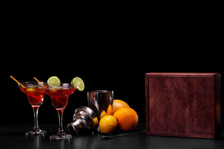 Close-up of two margarita glasses full of sweet, alcohol red cocktails with colorful straws and slices of lime on a black background. A heap of organic oranges, a box and shaker for drinks on a table.の写真素材