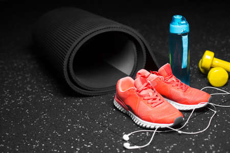 A close-up picture of a blue bottle for water, black stretching mat, bright pink training shoes, yellow equipment for physical exercises and modern phone with handphones on a spotted black background.の写真素材