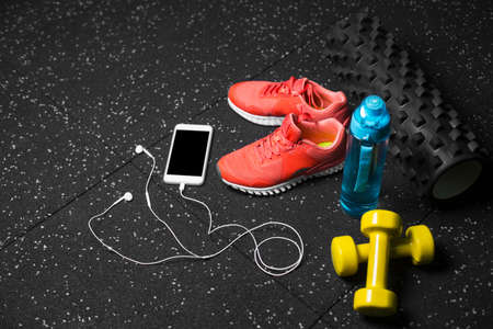 Blue sportive bottle, bright pink training shoes, yellow dumbbells, black pilates mat and smartphone on a black floor background. A colorful set of sportive accessories for gym training. Copy space.の写真素材