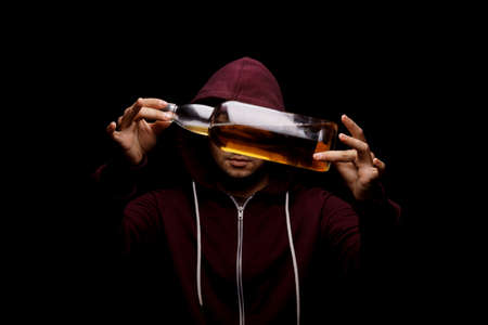 A handsome guy wearing a loose red hoodie with a big transparent bottle full of light alcoholic drink on a saturated black background. A desperate, lonely male with whiskey, beer or scotch in hands.の写真素材