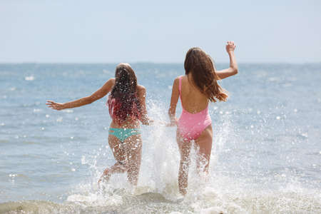Stunning and good-looking ladies wearing colorful swimwears splashing in a blue sea on a sunny sky background. Happy, handsome teenage girls having a good time for summer vacation. Copy space.の写真素材