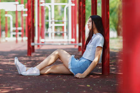 A full-length photo of a beautiful teenage girl in casual clothes and pink gumshoes sitting on an athletic field ground.の写真素材