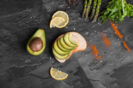 A sandwich with green slices of avocado sprinkled with seasonings, asparagus, salad leaves on a gray...