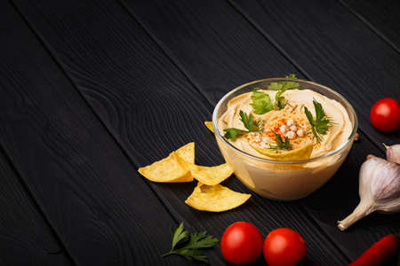 A glass bowl full of nutritious paprika hummus decorated with chickpea. Arabic dish surrounded by the tomatoes, garlic, and gold nachos on a black wooden background. Copy space. Food concept.の写真素材