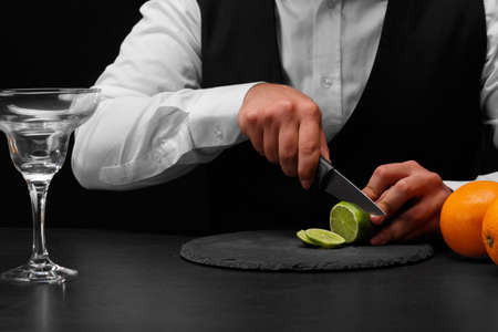 A bar counter with fresh vivid oranges, a glass for cockail, a bartender slices a sappy green lime with a knife on a black background. Night clubs, restaurant, cafe, party, concept.の写真素材