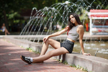 An appealing beautiful girl with long hair seating at the edge of the fountain on a natural blurred background.の写真素材