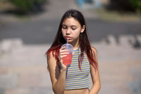 A beautiful girl with long hair drinking a fruit cocktail in a park on a natural blurred background.の写真素材