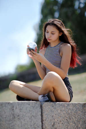 A beautiful girl with a cocktail and cell phone sitting on a stone in a park on a natural blurred background.の写真素材