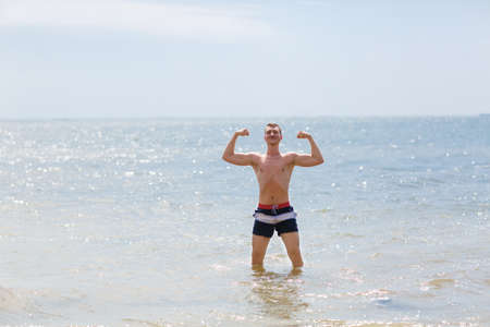 A strong young fellow in blue swimming trunks standing in a blue sea on a natural blurred background.の写真素材