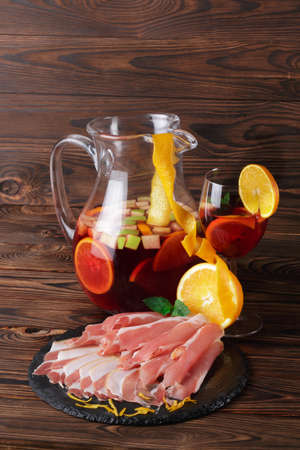 A wooden table of a glass mason jar full of refreshing beverage with slices of apple and oranges, a plate of prosciutto and ham on a wooden brown background.の写真素材