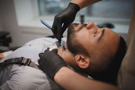 A muscular man sitting in a chair in a barber shop shaved by a barber on a light blurred background.の写真素材