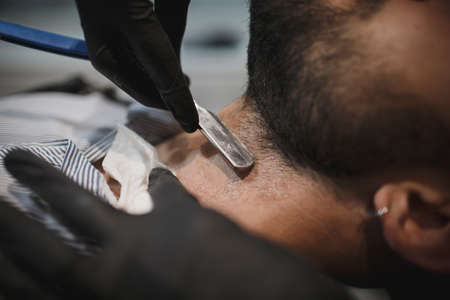 A muscular man sitting in a chair in a barber shop shaved by a barber on a light blurred background.の写真素材