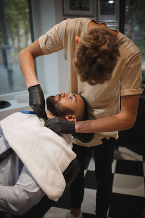 A handsome man in a barber chair is getting shaved by a barber with a razor on a blurred light background.の写真素材