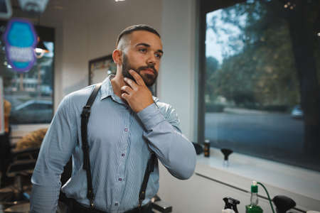 Bearded man on a blurred background. Handsome man touching new beard in a barbershop. Hipster style concept. Copy space.の写真素材