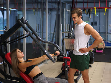 A sporty girl doing weight exercises on a simulator with the assistance of her personal trainer at...