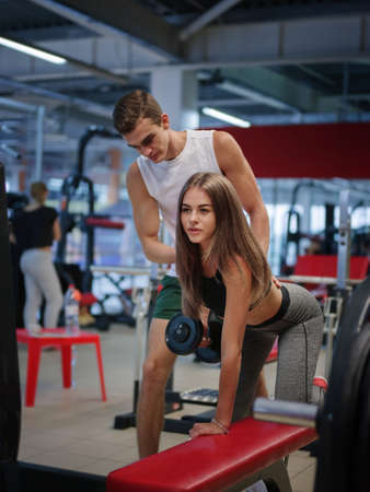 A sporty girl training with dumbbells on a colorful gym background. A coach helping her client in a fitness club.の写真素材