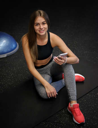 A pretty female wearing sports uniform with a phone on a gym background. Fitness, activity, and bodybuilding concept.の写真素材