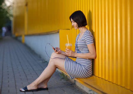 A beautiful girl with a phone and fresh juice in her hands sitting on a colorful street background. Outdoors concept.の写真素材