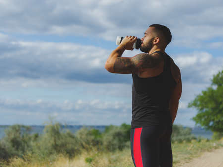 A strong bodybuilder with a muscular body and a tattoo drinking water after a workout on a natural blurred background.の写真素材