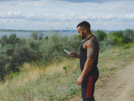 A muscular handsome bodybuilder with a tattoo on his shoulder listening music after a workout on a natural background.の写真素材