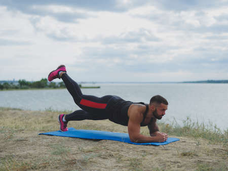 A strong tattooed bodybuilder standing in a plank on a bank of a river on a blurred natural background.の写真素材