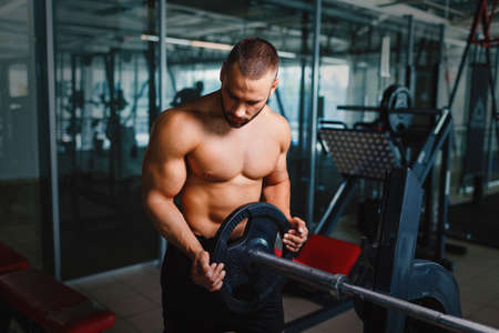 A sexy man with a muscular torso holding a barbell in a gym on a dark blurred background.の写真素材