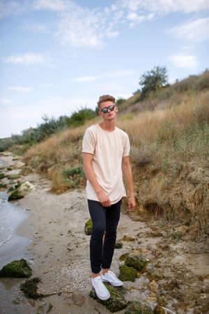 An attractive fellow in a white T-shirt and sunglasses standing on a riverbank on a blurred natural background.の写真素材