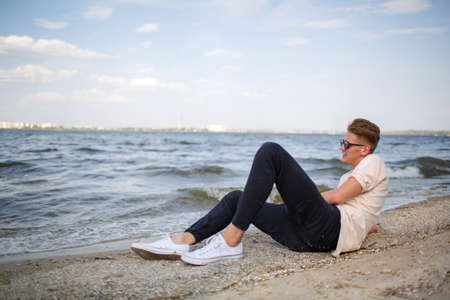 An attractive fellow in a white T-shirt sitting on a riverbank on a natural blurred background.