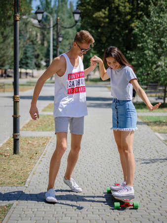 An attractive smiling fellow teaching a girl riding on a longboard in a park on a natural blurred...