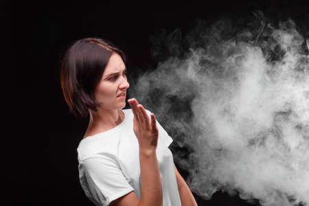 A young woman does not like the smoke of a cigarette or an electronic cigarette on a black background. Health concept.の写真素材