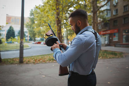 A confident handsome criminal holding a gun on an urban background. An elegant gangster in a blue...