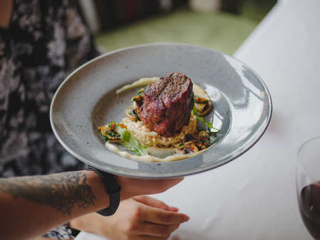 A close-up picture of a tattooed hand with a blue round plate full of an exquisite dish on a blurred background. Juicy meat in a plate. Expensive food. Restaurant, food, service concept. Copy space.の写真素材