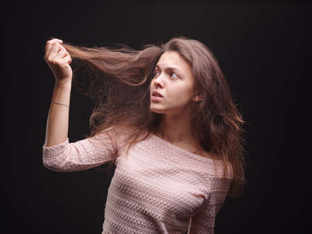 Pretty, worried lady touching hair on a black background. Young woman disappointed with her hair. Fragile hair concept.の写真素材