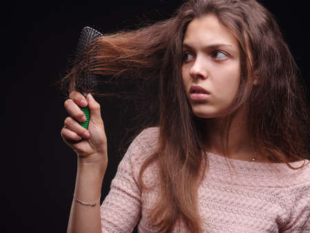 Woman brushing tangled hair with a comb on a black background. Girl looking at damaged sick hair. Hair problems concept.の写真素材