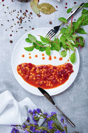 Top view of a gray table with white plate, canned beans, silver knife and fork, different fragrant seasonings, bay leaves and green salad leaves on a gray light background.の写真素材