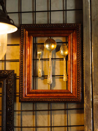 Shiny decorative spoons and silver fork in a brown wooden frame hanging on a cafe wall background. A set of antique utensil for bar or pub decoration. Luxurious ethnic spoons and forks.の写真素材