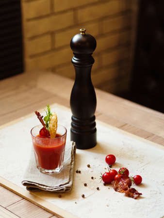 A top view of a glass of Bloody Mary and cherry tomatoes on a white fabric and on a wooden table. Stylish pepper mill and alcohol cocktail with bacon on a top of glass on a blurred background.の写真素材