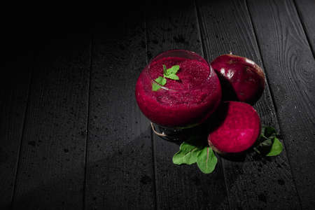 Close-up picture of a bright set of a spacious glass with a blended vegetable smoothie and claret-colored beet. Beet with leaves next to a healthful beverage on a black wooden table background.の写真素材