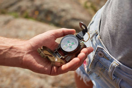 Hand with compass at mountain road at sunset sky. Searching direction with a compass in summer mountains. Close-up view of hand showing little round compass to camera, with incognito man.の写真素材
