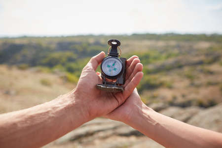 Hand with compass at mountain road at sunset sky. Searching direction with a compass in summer mountains. Close-up view of hand showing little round compass to camera, with incognito man.の写真素材