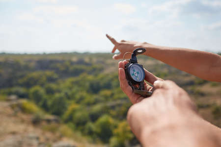 Tourists's hands with compass at mountain road at sunset sky. Close-up view of hand showing little round compass to camera, with incognito man. Point of view shot.の写真素材