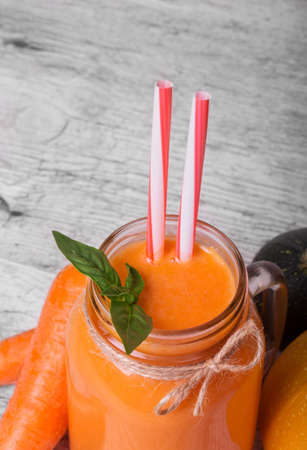 Closeup of a fresh carrot, a mason jar with refreshing orange smoothie with succulent green leaves of mint and striped drinking straw on a light blurred background.の写真素材
