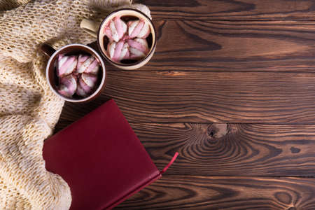 Coffee with marshmallows, woolen scarf and book on a wooden background. Winter hot chocolate. Coziness concept.の写真素材
