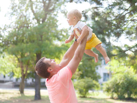 Cute little boy in hands of a happy dad on a park background. Dad playing with baby son concept. Copy space.の写真素材