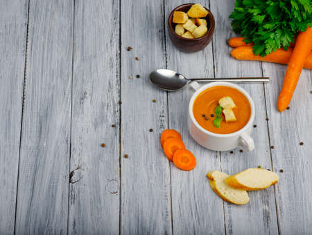 Curried carrot soup with cream fresh herbs and other vegetables on wooden grey table. Two piese of white bread big orange carrots. Close-up of plate with soup. Copy space. Food concept.の写真素材