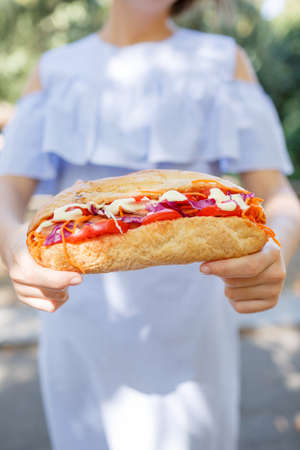 Beautiful young woman with sunny smile holding juicy burger and soda. Close-up of fast food on the nature background.の写真素材
